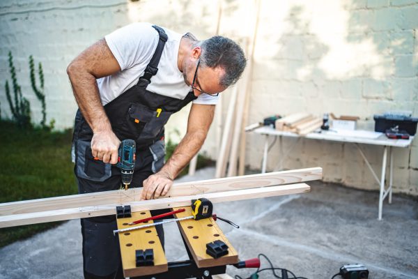 Mature male carpenter using electric drill on a wooden plank outdoors.